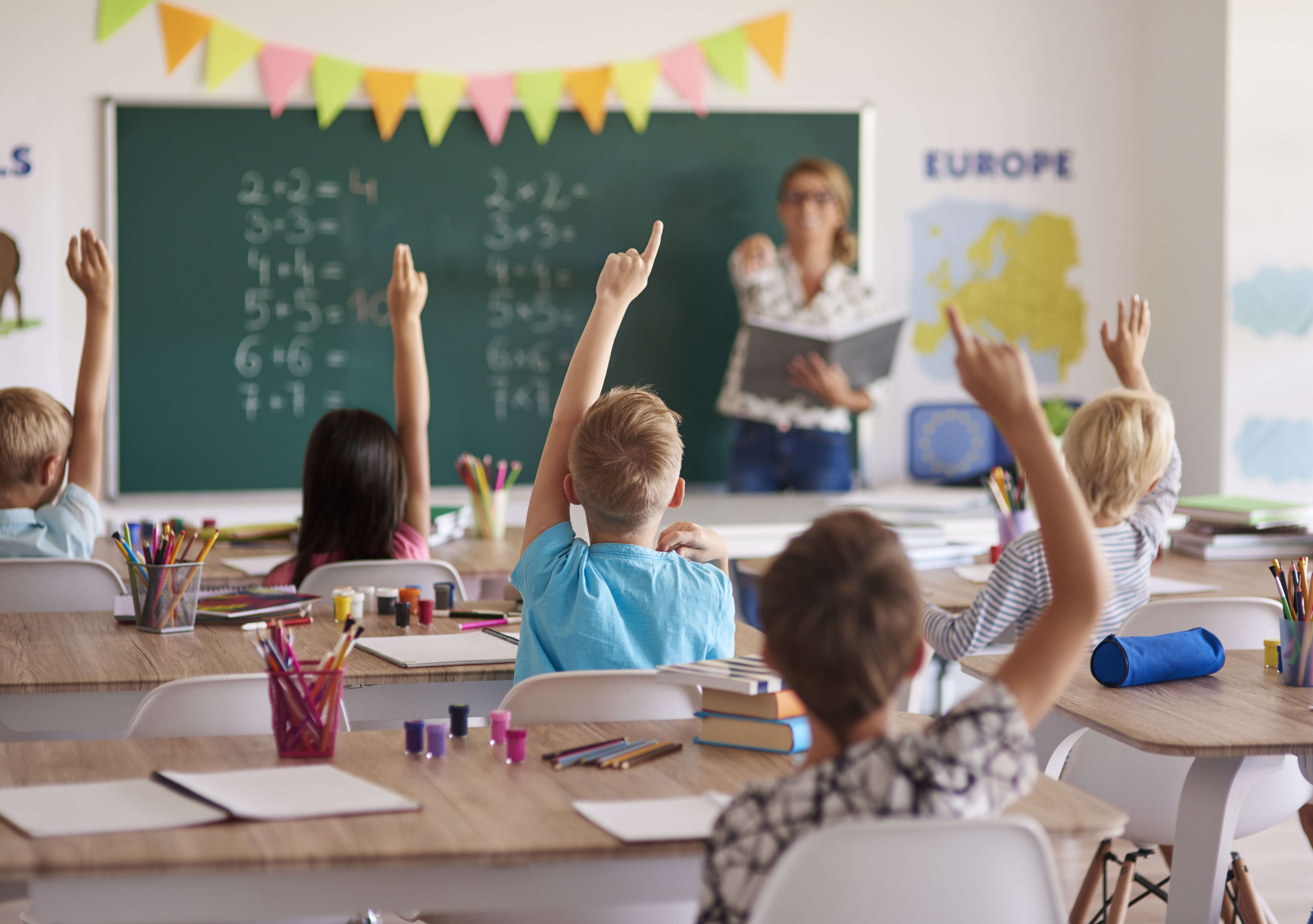Žiaci v triede sedia pri laviciach a s nadšením dvíhajú ruky, aby odpovedali na otázku učiteľky, ktorá stojí pri tabuli s matematickými príkladmi. V triede sú aj školské pomôcky a mapy.