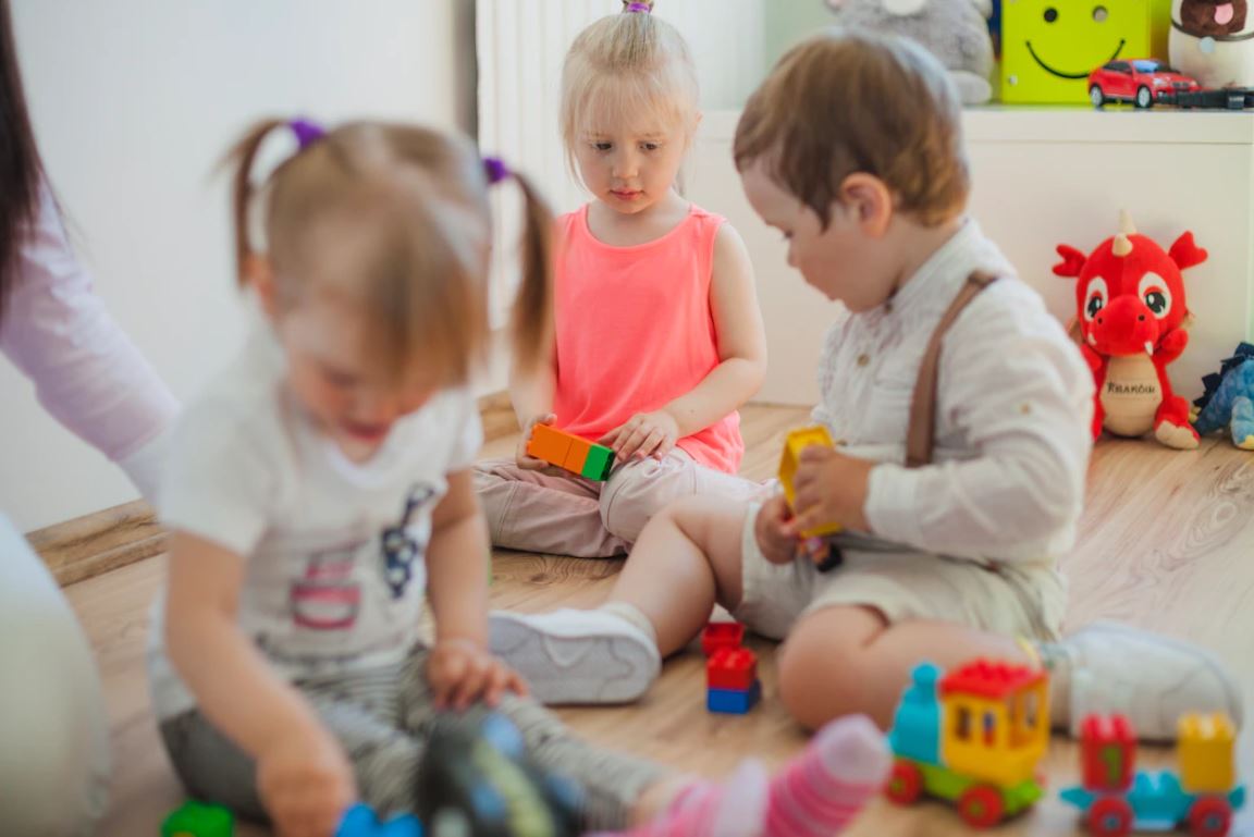 Tri malé deti sedia na podlahe v interiéri a hrajú sa s farebnými stavebnicami a hračkami. V pozadí je vidieť plyšového draka a ďalšie detské predmety.