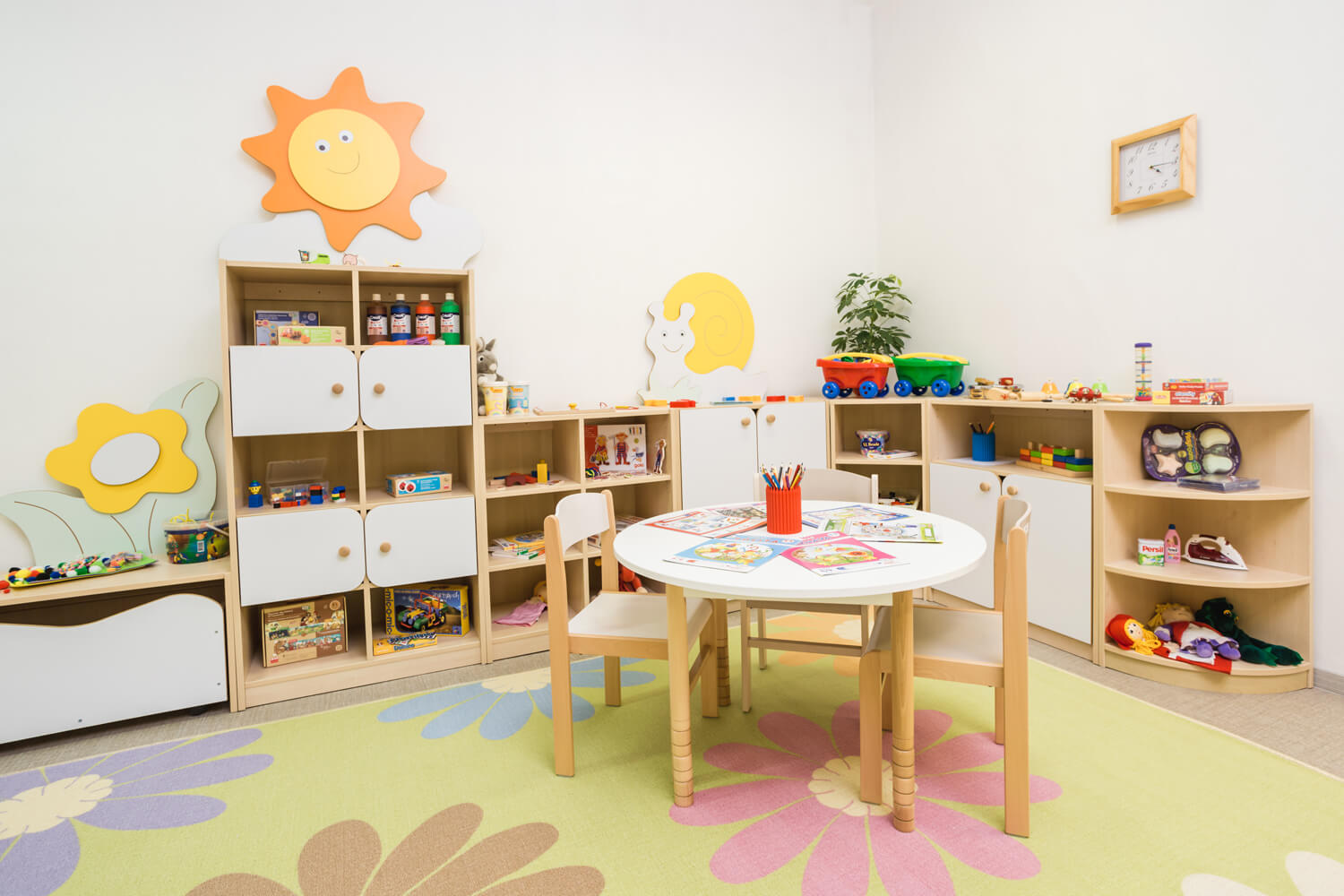 Třída mateřské školy s dřevěným stolem, židlemi a úložnými skříňkami, plná kreativních pomůcek a hraček pro děti.
