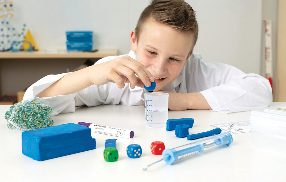Chlapec v laboratórnom plášti vykonáva vedecký experiment s odmerným valcom, kockami a ďalšími edukačnými pomôckami.