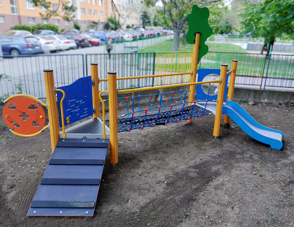Újonnan telepített játszótéri mászóka csúszdával, mászóráccsal és interaktív elemekkel. A háttérben kerítés és parkoló látható.