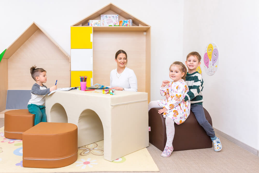 Deti s učiteľkou v školke sediaci na molitanových sedačkách so sériou Mesto v pozadí.